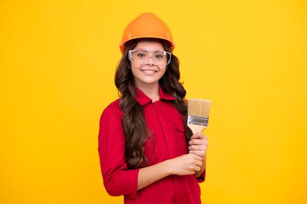 Concept de rénovation et de construction pour enfants Petite fille dans un casque de protection et un pinceau pour peindre des murs isolés sur fond jaune Visage heureux émotions positives et souriantes d'une adolescente