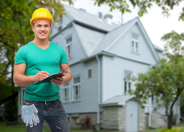 concept de réparation, de construction, de personnes, de construction et d'entretien - ouvrier masculin souriant dans un casque de protection prenant des notes dans le presse-papiers sur fond de maison