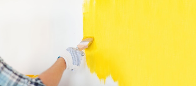 concept de réparation et de rénovation domiciliaire - gros plan d'une main masculine dans des gants peignant un mur avec de la peinture jaune