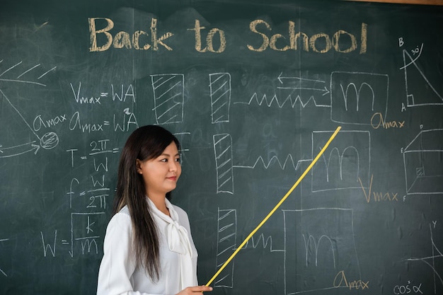 Concept de retour à l'école. Enseignante asiatique souriant avec un bâton en bois pointant vers le tableau noir à l'école dans la salle de classe, Heureuse belle jeune femme debout tenir le pointeur vers le panneau arrière