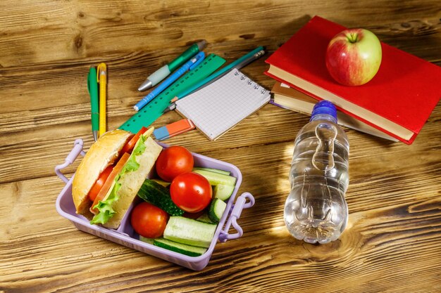 Concept de retour à l'école Fournitures scolaires livres bouteille d'eau pomme et boîte à lunch avec des hamburgers et des légumes frais sur une table en bois