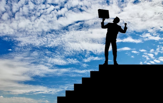 Concept de réussite commerciale. Silhouette d'un homme d'affaires heureux avec une mallette dans ses mains à la hauteur des marches sur fond de ciel