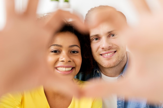 Photo concept de saint valentin, d'amour et de personnes - heureux couple multiethnique regardant à travers la forme de coeur faite de doigts