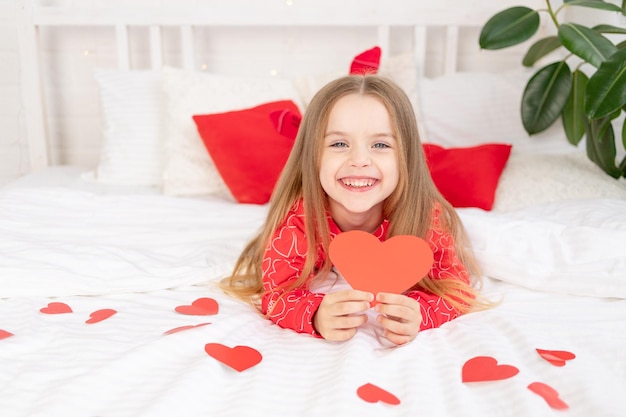 Le concept de la Saint-Valentin une douce enfant fille sur un lit blanc à la maison en pyjama rouge parmi les coeurs tenant un coeur de la Saint-Valentin dans ses mains souriant de bonheur félicitant pour les vacances