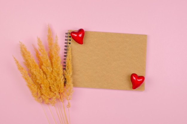 Le concept de la Saint-Valentin fête des mères Bloc-notes vierge avec du papier kraft sur fond rose avec des coeurs et de l'herbe de la pampa Formulaire de félicitations ou d'invitations