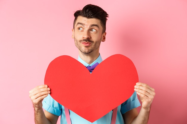 Concept de la Saint-Valentin. Homme romantique de rêve à gauche et montrant une grande découpe de coeur rouge, debout sur rose