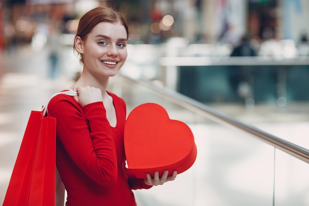 Concept de la Saint-Valentin. Jeune femme avec sac à provisions et coffret cadeau en forme de coeur rouge portrait au magasin du centre commercial