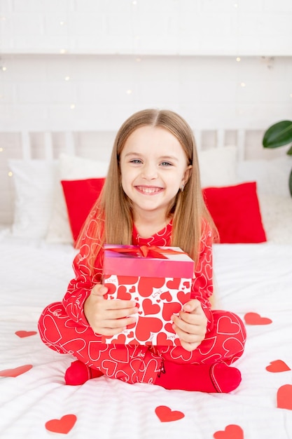 Le concept de la Saint-Valentin, une jolie petite fille est assise sur le lit à la maison en pyjama rouge et tient un cadeau dans ses mains et sourit ou rit de bonheur en félicitant les vacances