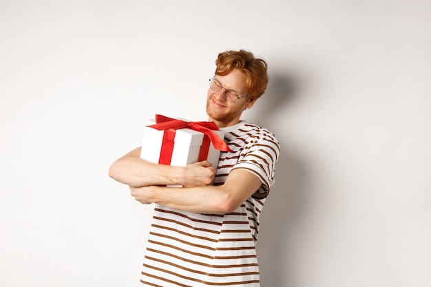 Concept de Saint Valentin et vacances. Heureux homme rousse étreignant sa boîte-cadeau et souriant ravi, debout sur fond blanc.