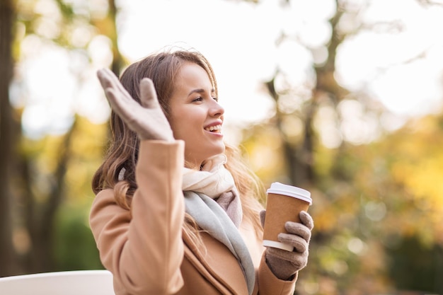 concept de saison, de boissons chaudes et de personnes - belle jeune femme heureuse buvant du café ou du thé dans une tasse en papier jetable et agitant la main dans le parc d'automne