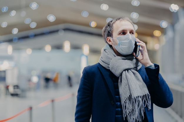 Concept de santé, de communication et d'épidémie. Un homme sérieux porte un masque médical de protection, se soucie de la sécurité de ne pas attraper le coronavirus, se promène dans un centre commercial, parle par téléphone portable. Protection antivirus