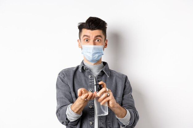 Concept de santé, de covid et de pandémie. Mec moderne en masque facial nettoyer les mains avec un antiseptique, à l'aide d'une bouteille de désinfectant, debout sur un fond blanc.