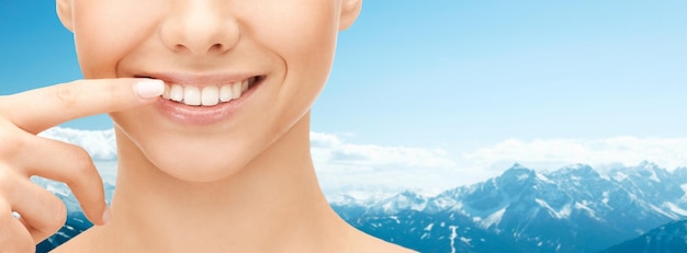 concept de santé dentaire, de beauté, d'hygiène et de personnes - gros plan d'un visage de femme souriant pointant vers les dents sur fond de montagnes bleues