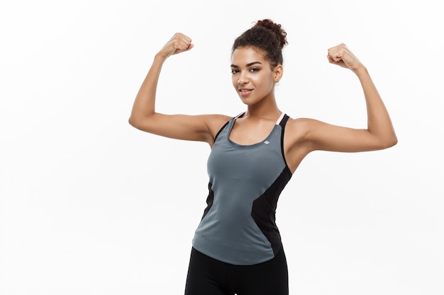 Concept De Santé Et De Remise En Forme - Portrait De Jeune Belle Afro-américaine Montrant Son Fort Muscle Avec Une Expression Faciale Gaie Et Confiante. Isolé Sur Fond De Studio Blanc.