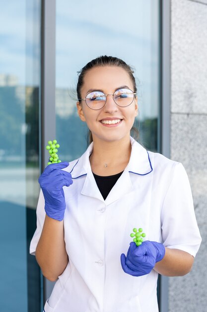 Photo concept scientifique, chimie, biologie et médecine - femme scientifique européenne heureuse en blouse blanche, lunettes et gants en caoutchouc montrant des molécules vertes et regardant la caméra à l'extérieur