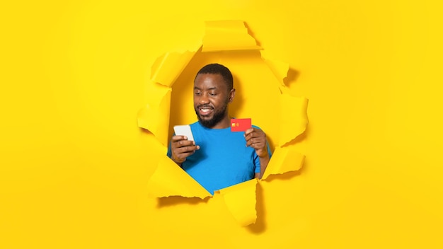 Concept de shopping mobile Homme noir utilisant une carte de crédit et un smartphone posant dans un trou de panorama de papier jaune déchiré