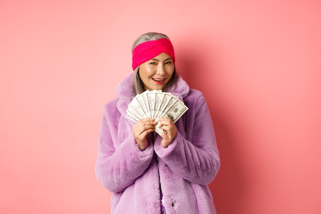 Concept de shopping et de mode. Femme âgée asiatique à la mode pensant à l'achat de nouveaux vêtements, montrant de l'argent en dollars et souriant fond rose gourmand