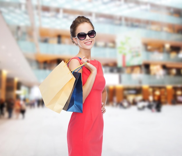 concept shopping, vente, noël et vacances - femme élégante souriante en robe rouge et lunettes de soleil avec des sacs à provisions