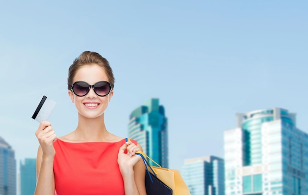 concept shopping, vente, noël et vacances - femme élégante souriante en robe rouge avec des sacs à provisions et une carte en plastique