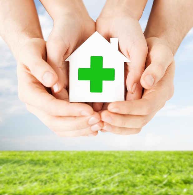 Photo concept de soins, d'aide, de charité et de personnes - gros plan de mains tenant une maison en papier blanc avec un signe de croix verte sur fond de ciel bleu et d'herbe