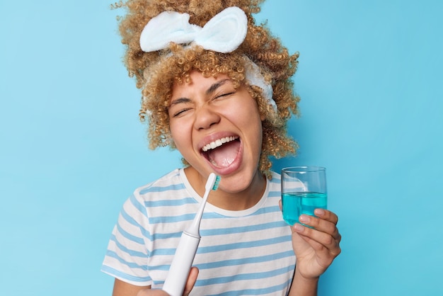 Concept de soins dentaires Une jeune femme positive aux cheveux bouclés et peignés tient une brosse à dents électrique et un verre de rince-bouche chante et s'amuse pendant la routine du matin porte un bandeau t-shirt rayé décontracté