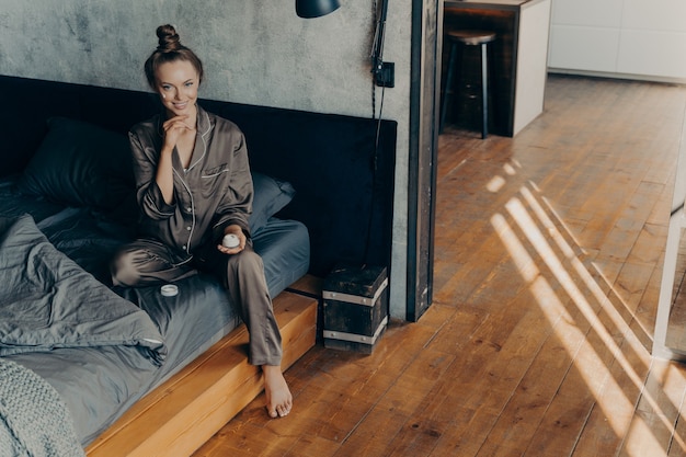 Concept de soins de la peau. Belle jeune femme souriante appliquant une crème hydratante pour le visage pendant les procédures cosmétiques quotidiennes alors qu'elle était assise sur le lit après s'être réveillée tôt le matin en pyjama en satin