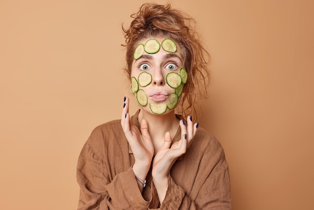 Concept de soins de la peau et de bien-être Une femme surprise a les cheveux en désordre applique des tranches de concombre sur le visage pour hydrater vêtue de vêtements de nuit a les yeux largement ouverts isolés sur fond de studio beige