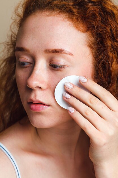 Concept de soins de la peau chez les adolescentes Jeune fille rousse aux cheveux bouclés nettoie son visage avec un coton