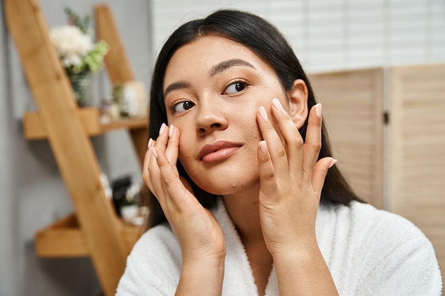 concept de soins de la peau jeune femme asiatique en robe de bain appliquant de la crème hydratante sur le visage et détournant le regard