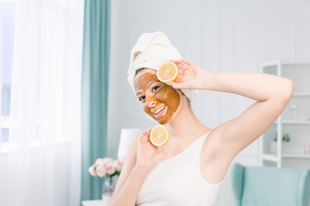 Concept de soins de la peau de procédures de beauté. Jeune femme avec un masque d'argile de boue du visage brun sur son visage dans la salle de bain, avec une serviette blanche sur la tête, tenant des demi-citrons