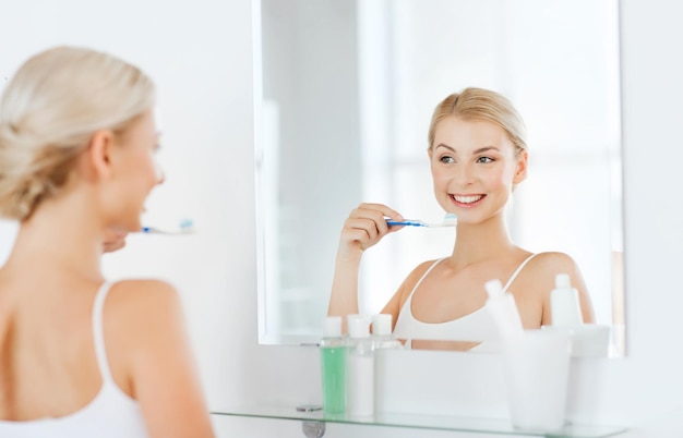 concept de soins de santé, d'hygiène dentaire, de personnes et de beauté - jeune femme souriante avec une brosse à dents nettoyant les dents et regardant au miroir dans la salle de bain à la maison