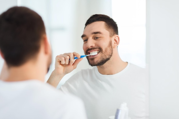 concept de soins de santé, d'hygiène dentaire, de personnes et de beauté - jeune homme souriant avec une brosse à dents nettoyant les dents et regardant au miroir dans la salle de bain à la maison