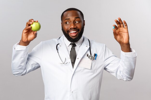 Concept de soins de santé, de médecine et de mode de vie sain. Sympathique médecin afro-américain en blouse blanche, tenant une pomme et une pilule, suggère aux patients de rester en bonne santé sans médicament, de manger plus de fruits