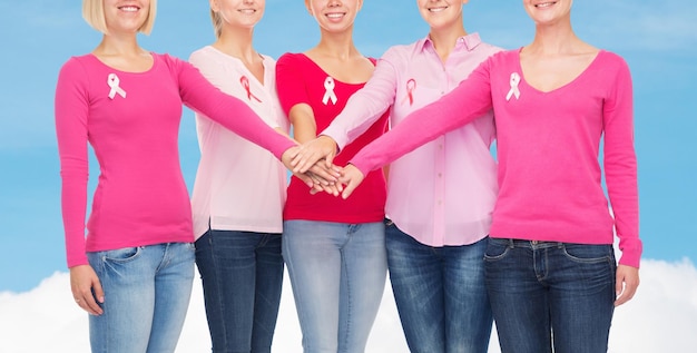 concept de soins de santé, de personnes, de gestes et de médecine - gros plan de femmes souriantes en chemises vierges avec des rubans roses de sensibilisation au cancer du sein mettant les mains sur le ciel bleu et fond de nuage blanc