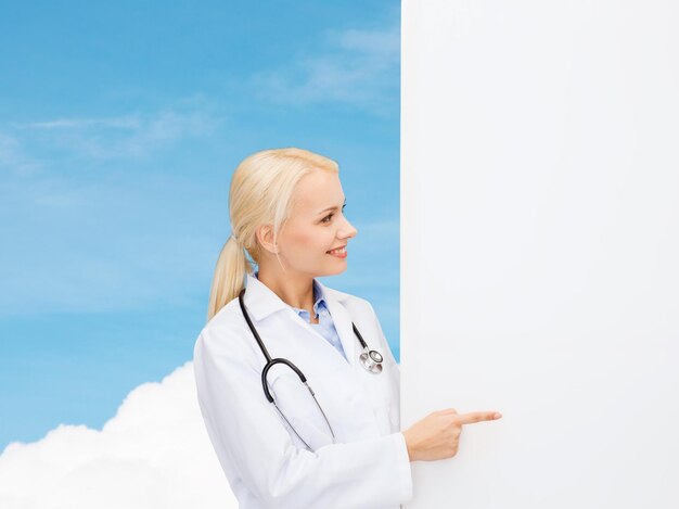 concept de soins de santé, de publicité, de personnes et de médecine - femme médecin souriante avec stéthoscope montrant quelque chose sur fond de ciel bleu