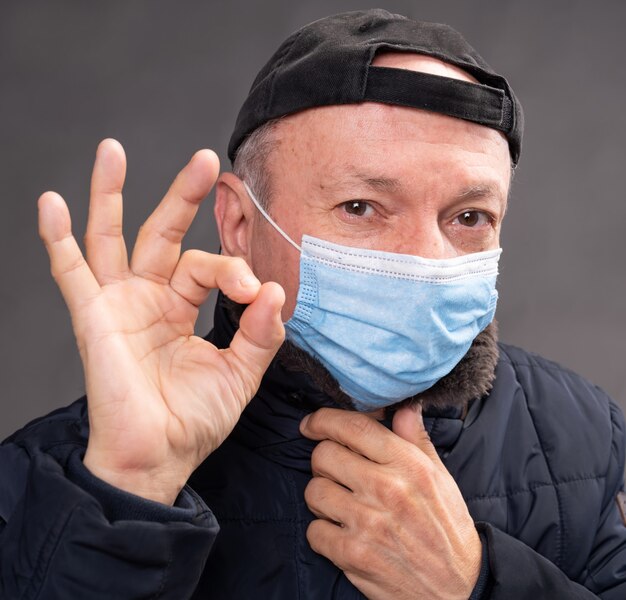 Concept de soins de santé. Senior homme en masque de protection qui pose en studio sur fond gris. Gestes OK signe