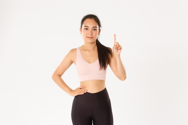 Concept de sport, bien-être et mode de vie actif. Souriant instructeur de fitness asiatique, fille en cours d'enseignement de vêtements de sport, montrant la règle numéro un, lever le doigt et sourire narquois, debout mur blanc.