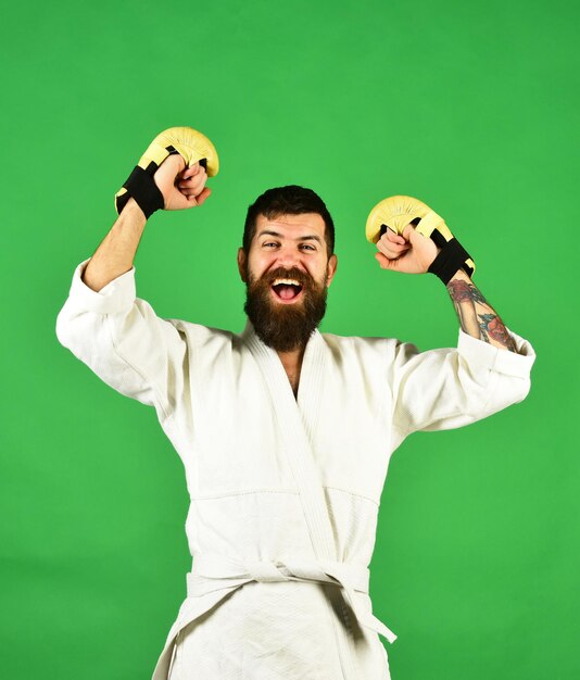 Concept de sport et de combat. Homme avec un visage et une barbe gagnants excités sur fond vert. Guy pose en kimono blanc portant des gants de boxe dorés. Un combattant de MMA remporte une compétition d'arts martiaux