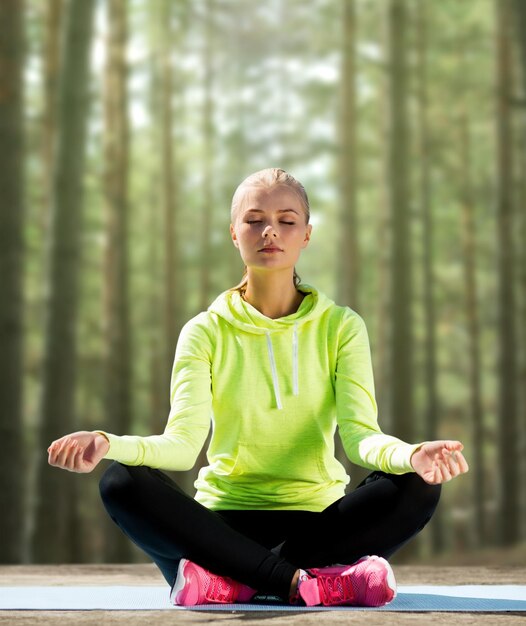 concept de sport, de fitness, de yoga et de personnes - jeune femme heureuse méditant dans la pose de lotus et assise sur un tapis sur fond de bois