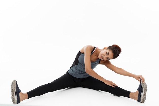 Concept de sport, de formation, de mode de vie et de remise en forme - portrait de femme afro-américaine heureuse qui s'étend de la jambe en position assise Isolé sur blanc.