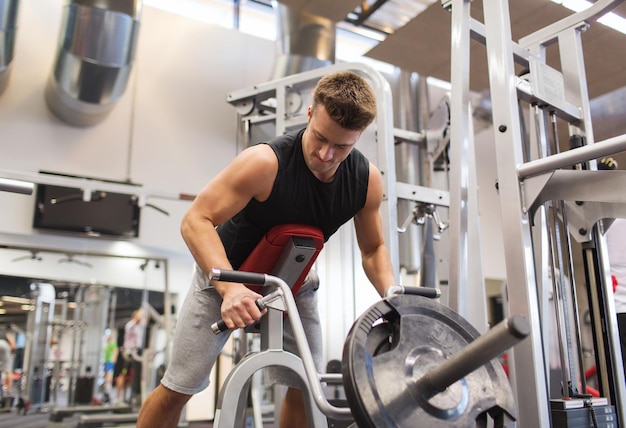 concept de sport, de musculation, d'équipement et de personnes - jeune homme avec des muscles de flexion d'haltères sur une machine à barre en t dans la salle de sport