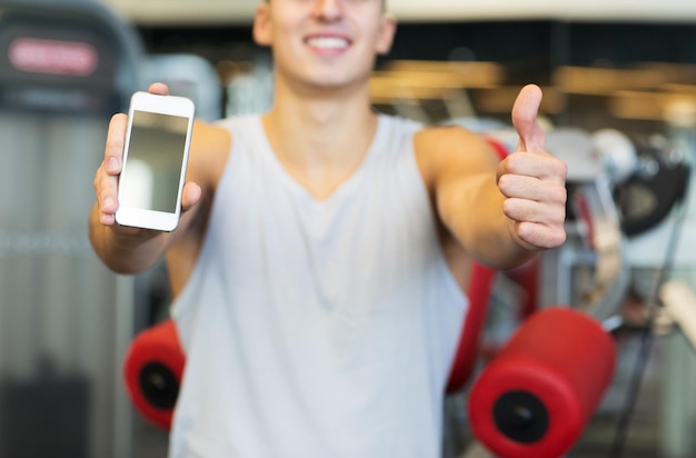 concept de sport, de musculation, de mode de vie, de technologie et de personnes - gros plan sur un jeune homme heureux montrant les pouces vers le haut et un écran vide de smartphone dans la salle de sport