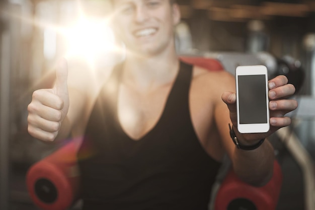 concept de sport, de musculation, de mode de vie, de technologie et de personnes - jeune homme heureux avec un smartphone montrant les pouces vers le haut dans la salle de sport