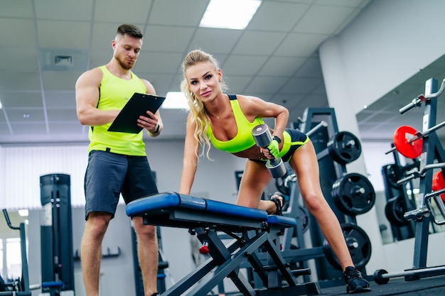 Concept de sport, de musculation, de style de vie et de personnes - jeune homme et femme avec haltères fléchissant les muscles et faisant des squats d'épaule dans la salle de sport. L'entraîneur et la fille de remise en forme s'entraînent au gymnase.