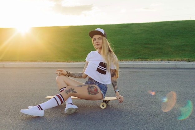 Concept de sport, de style de vie, d'extrême et de personnes - Tatouages de belles filles faisant du longboard sur la route de la ville par temps ensoleillé. Portrait hipster girl smiling avec un longboard au coucher du soleil.