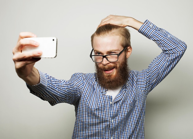 Concept de style de vie : un jeune homme avec une barbe en chemise tenant un téléphone portable et faisant une photo de lui-même en se tenant debout sur fond gris.