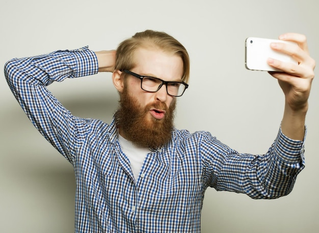 Concept de style de vie un jeune homme avec une barbe en chemise tenant un téléphone portable et faisant une photo de lui-même en se tenant debout sur fond gris