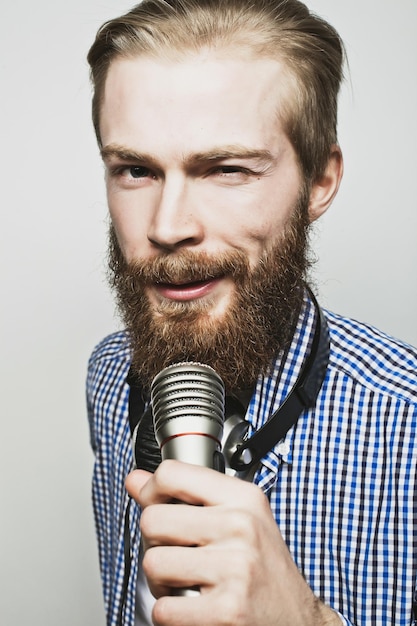 Concept de style de vie : un jeune homme avec une barbe portant une chemise blanche tenant un microphone et chantant. Sur fond gris. Photos tonifiantes spéciales à la mode.