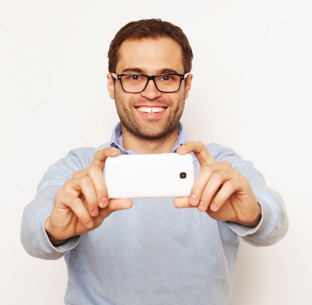 Concept de style de vie, de technologie et de personnes : un jeune homme en chemise tenant un téléphone portable et faisant une photo de lui-même en se tenant debout sur fond blanc.