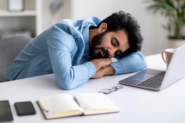 Concept de surmenage fatigué jeune homme indien faisant la sieste au bureau avec ordinateur portable